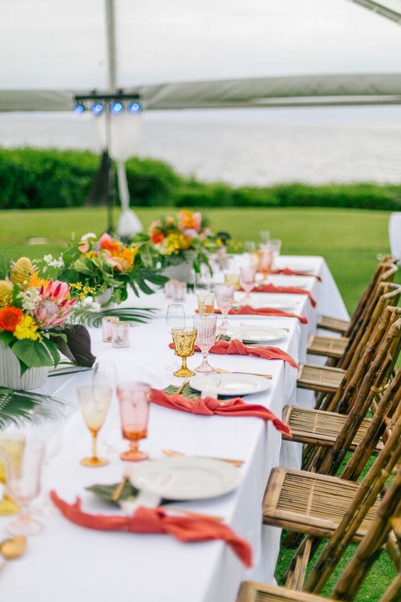 a bold and chic tropical wedding tablescape with bold blooms, pink napkins, colored glasses and tropical leaves
