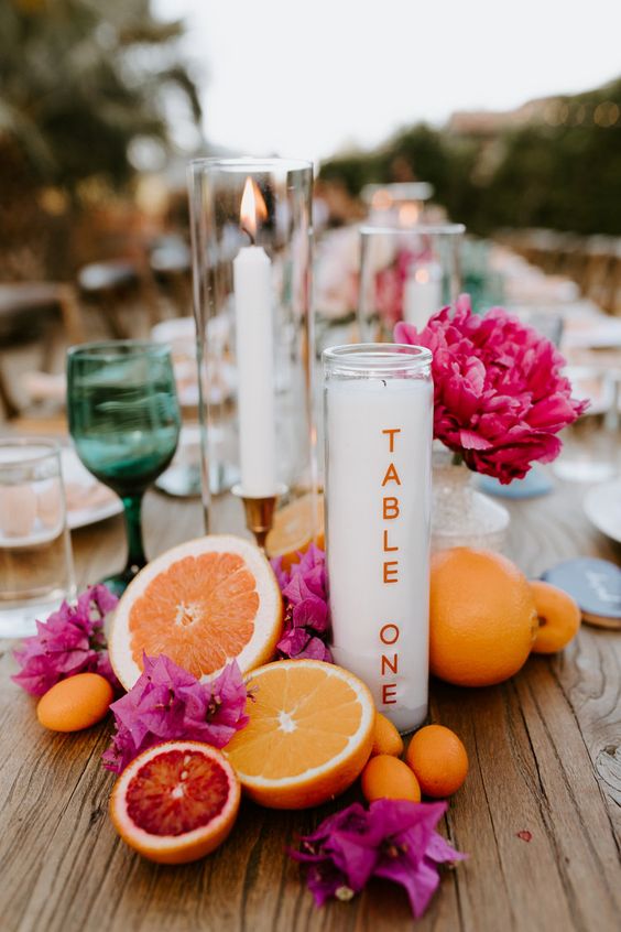 a bright tropical wedding centerpiece of citrus and bright fuchsia blooms, various candles and colored glasses is a gorgeous idea