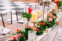 a bright tropical wedding tablescape with a neutral fabric and a greenery and bold floral runner, candles, neutral napkins and large tropical leaves