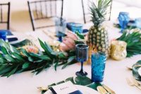 a bright wedding tablescape with a greenery and pink bloom runner, pineapples, navy glasses and napkins