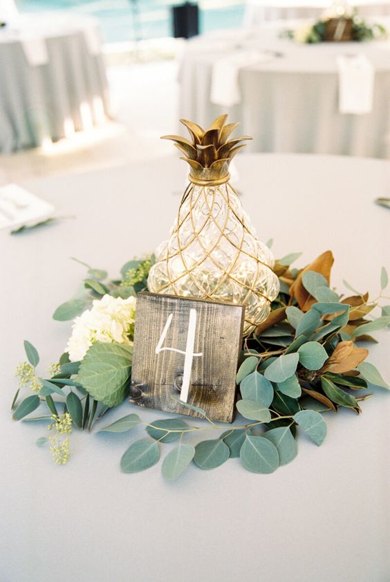 a chic wedding centerpiece of foliage, white blooms, a wooden plaque and a pineapple-shaped lantern with lights inside is awesome