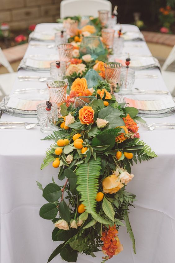 a colorful table runner with textural greenery and leaves, orange and rust flowers and kumquats is ideal for a tropical wedding