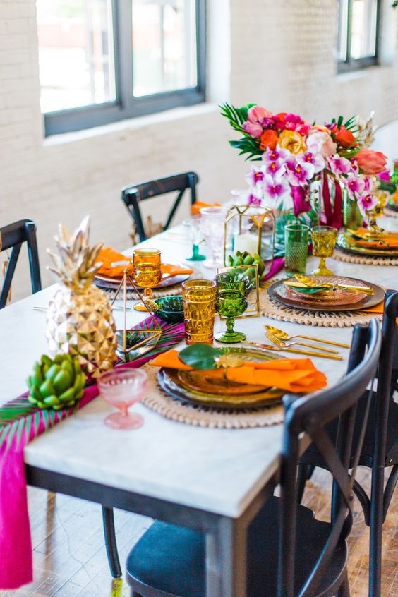 a colorful tropical boho wedding tablescape with colorful glasses, gold pineapples, gold candle lanterns and super bright blooms