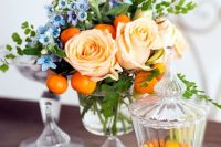 a colorful wedding centerpiece of a crystal bowl, with peachy roses, kumquats, blue blooms and greenery is a very cool and fun idea to rock