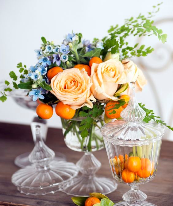 a colorful wedding centerpiece of a crystal bowl, with peachy roses, kumquats, blue blooms and greenery is a very cool and fun idea to rock