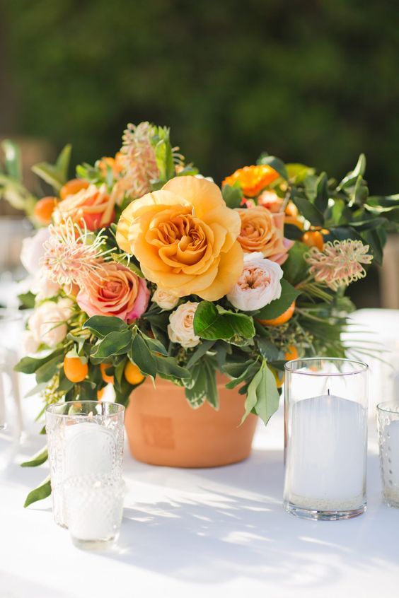 a colorful wedding centerpiece of a terra cotta pot, greenery, orange, pink and blush blooms and kumquats is amazing for spring or summer