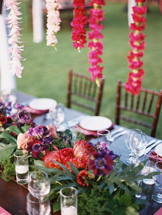 a colorful wedding table with a very bold floral centerpiece and garlands of blooms hanging overhead, some greenery and leaves and candles