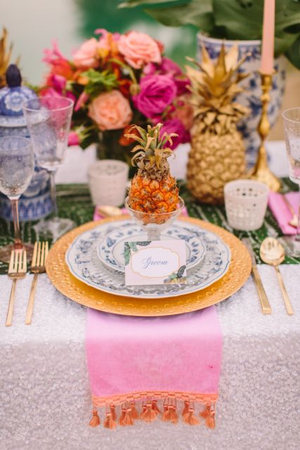 a colorful wedding tablescape with a pink napkin, gold chargers and cutlery, pineapples on the table and bold blooms