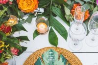 a colorful wedding tablescape with bold blooms and greenery, candles, woven chargers and tropical leaf menus