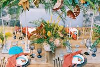 a colorful wedding tablescape with greenery, leaves and yellow blooms, blue stationery and red napkins