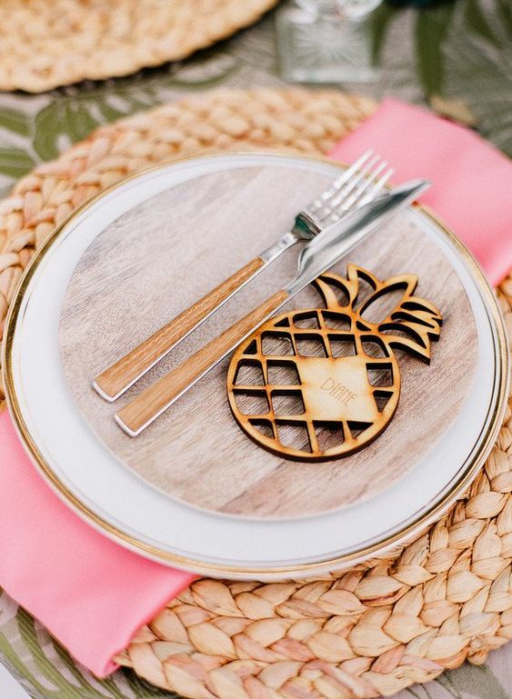 a creative and bright wedding table setting with a woven charger, neutral and wooden plates, pink napkins and a wood burnt pineapple