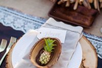 a creative wedding place setting with a wood slice charger, neutral porcelain and napkins and a mini pineapple in a wooden shell