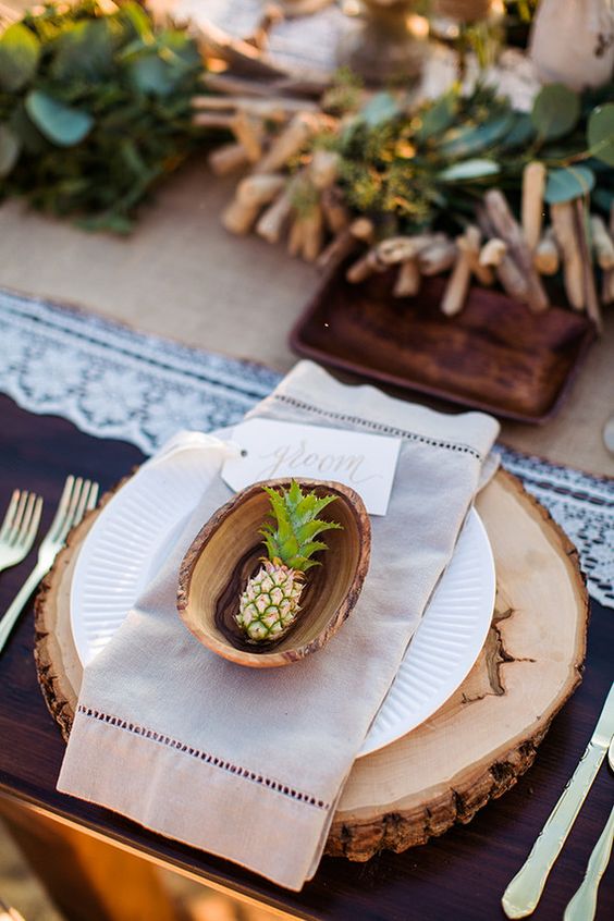 a creative wedding place setting with a wood slice charger, neutral porcelain and napkins and a mini pineapple in a wooden shell