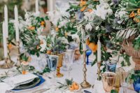 a fantastic Mediterranean wedding tablescape with green placemats and blue and white plates, with lush greenery and white and orange blooms and kumquats, a lace tablecloth