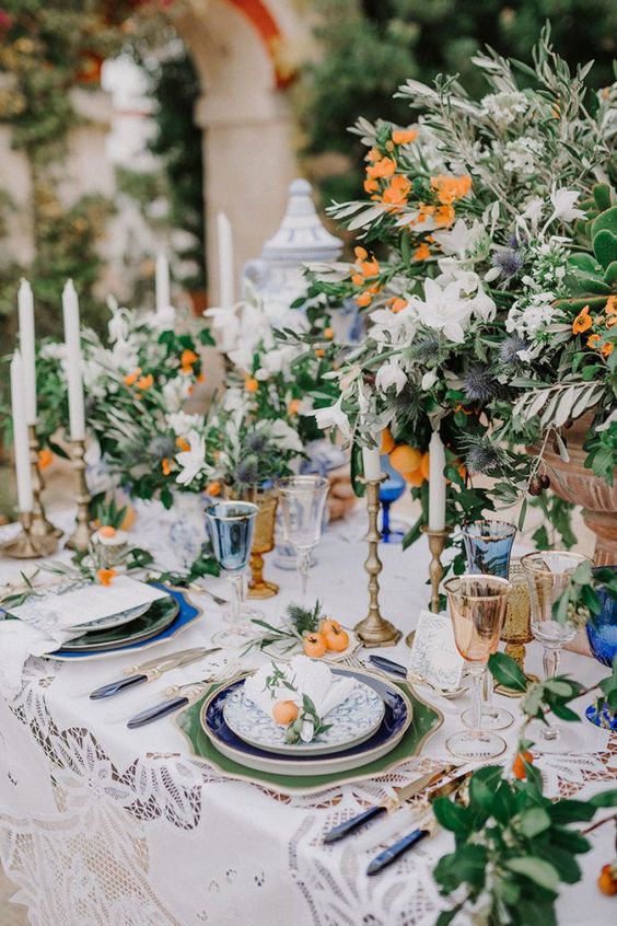 a fantastic Mediterranean wedding tablescape with green placemats and blue and white plates, with lush greenery and white and orange blooms and kumquats, a lace tablecloth