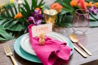 a fun bright tropical wedding table with tropical leaves, blue chargers, a pink napkin, gold candleholders and a small pineapple