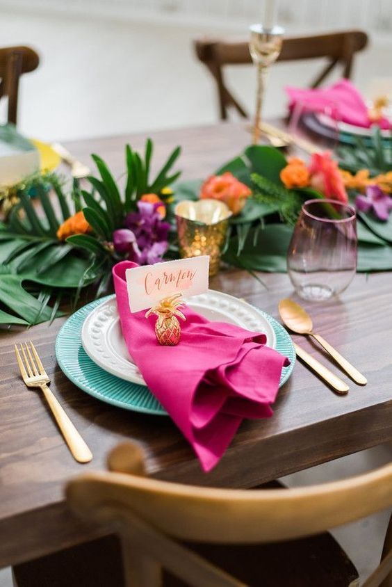 a fun bright tropical wedding table with tropical leaves, blue chargers, a pink napkin, gold candleholders and a small pineapple