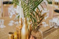 a glam metallic wedding centerpiece of a gilded pineapple, gold and copper vases and bottles with leaves and a gold candleholder