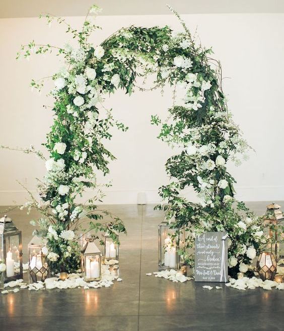 a lush wedding arch decorated with greenery and white blooms and surrounded with candle lanterns