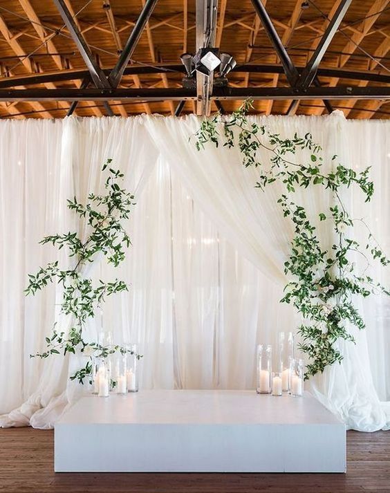 a minimalist wedding altar - a white platform, candles, greenery hanging on curtains