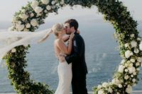 a round wedding arch decorated with greenery and white blooms plus a sea view for a breathtaking wedding ceremony
