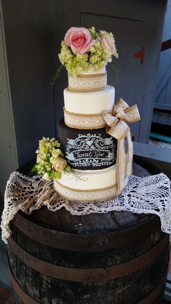 a rustic wedding cake with white and a chalkboard tier, with burlap and lace ribbons, with chalking to personalize the cake and some blooms on top