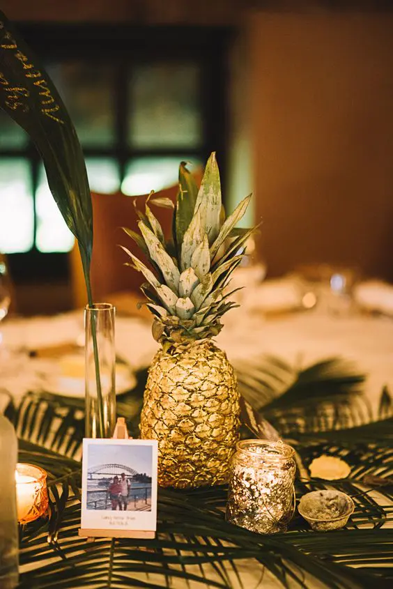a simple wedding centerpiece of tropical foliage, a gilded pineapple, a bud vase with a leaf, a gilded candleholder and some photos