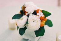 a small and cool wedding centerpiece of a white vase, with white ranunculus, leaves and kumquats is a pretty and bright solution