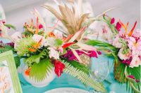 a super bold wedding tablescape with a turquoise tablecloth, a pink fan, bright blooms and leaves and a sign in a green frame