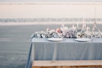 a blue beach wedding tablescape with a blue tablecloth, seashells, corals and white candles looks like it’s out of the sea