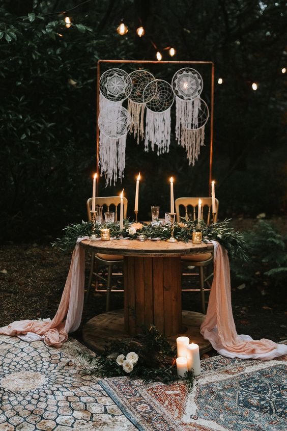 a boho woodland reception table with a greenery runner, a pink one, candles, gilded candleholders and an arch with macrame