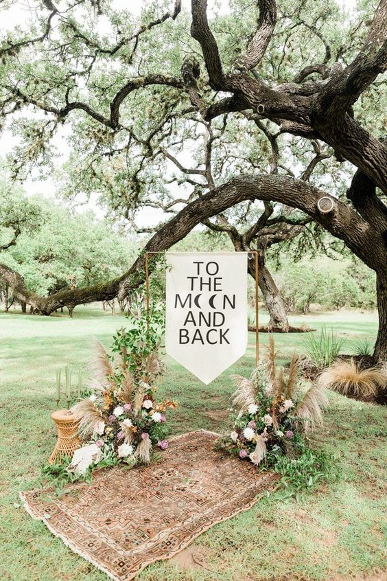 a boho woodland wedding arch with a banner, greenery, pampas grass and neutral blooms and a boho rug is beautiful