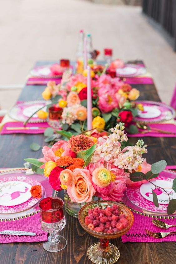 a bright and colorful table setting with hot pink placemats, bright blooms, pink candles, berries and colorful glasses