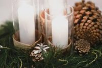 a chic and simple winter wedding centerpiece of evergreens, pinecones, pillar candles in glasses wrapped with bark