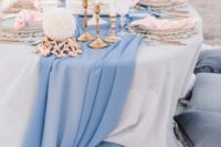a chic beach wedding tablescape with a blue runner, pink napkins, a naked wedding cake topped with blooms and white candles