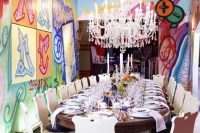a colorful wedding reception space with bold graffiti walls, a long dining table, refined chairs, a beautiful chandelier and white linens and candles