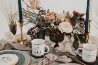 a cool winter mountain wedding table with a lush floral centerpiece, teal candles, plates and metal mugs and a embroidery hoops with mountains