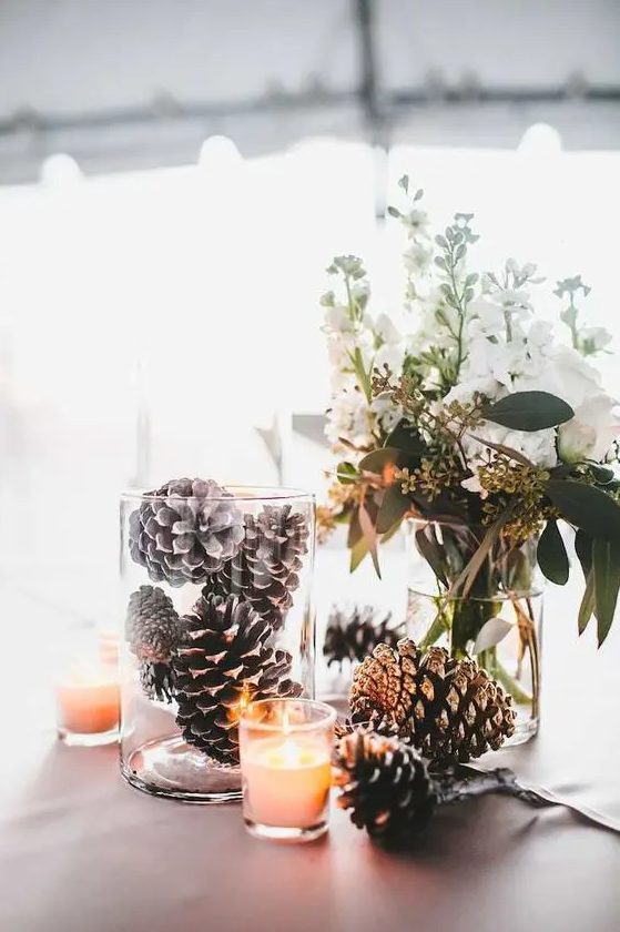 a creative winter wedding centerpiece of white blooms and greenery, pinecones in a glass and more on the table, some candles is cool