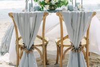 a light blue beach wedding tablescape with light blue candles, glasses, plates and linens, neutral blooms and driftwood