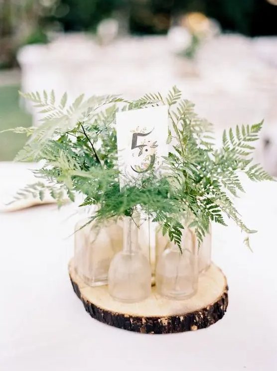 a pretty cluster woodland wedding centerpiece of a wood slice, bottles and fern leaves and a table number is chic