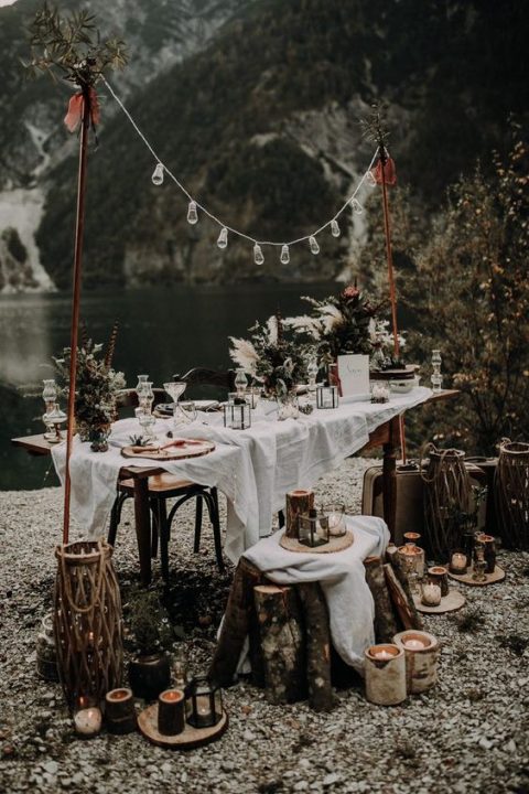 a rustic boho reception space by the lake with candles, bulbs and textural greenery decor