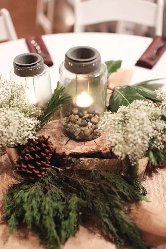 a rustic wedding centerpiece with mason jars, pebbles, a wood slice, evergreens, baby's breath
