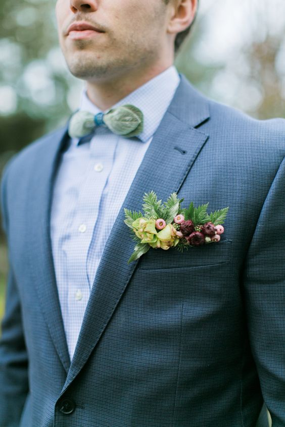 a stylish woodland groom's look with a grey suit, a light blue shirt, a green bow tie shaped as leaves, a burgundy and yellow pocket square with blooms and berries