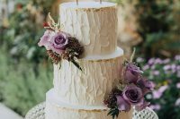 a textural neutral wedding cake with gold leaf, purple blooms and greenery and a calligraphy topper