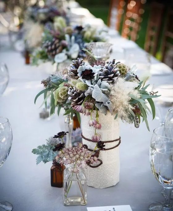 a vase wrapped with book pages, pale leaves, pinecones and textural herbs for a unique snowy-inspired centerpiece