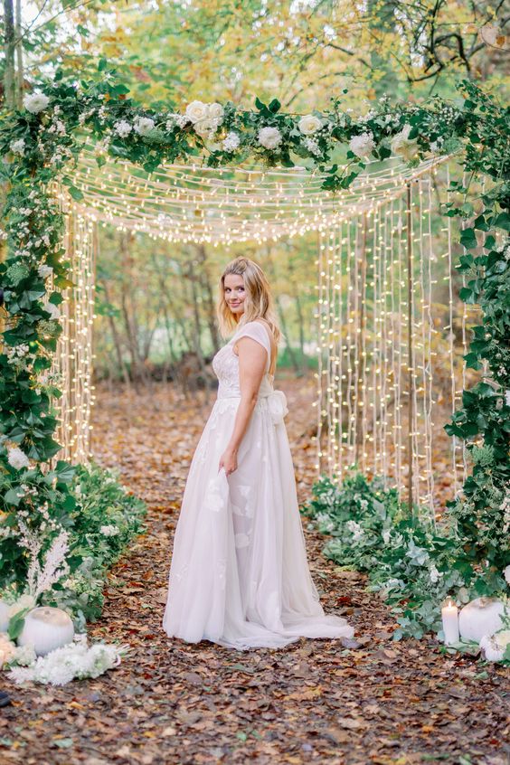 a woodland wedding arch of lights, greenery and white blooms and candles on the ground looks magical and enchanting