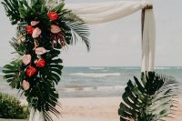 a chic tropical wedding arch of bamboo with white fabric, monstera leaves, pink and red flowers