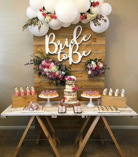 a cool dessert table with a wooden backdrop with greenery, blooms and white balloons and lots of desserts