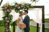 a cool tropical wedding arch in dark wood, with airy fabric, tropical leaves and bright blooms and king proteas