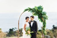 a curved wedding arch decorated with tropical leaves only for a modern tropical wedding